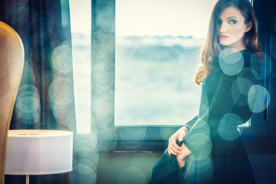 Portrait of a woman seated beside a window, with long dark hair and dressed in black.
