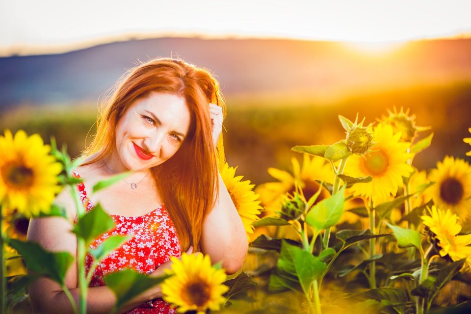A smiling woman with long reddish hair and a patterned red dress is pictured among flowering sunflowers.