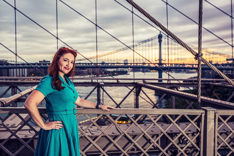 A smiling woman in a turquoise dress leans on a bridge railing.