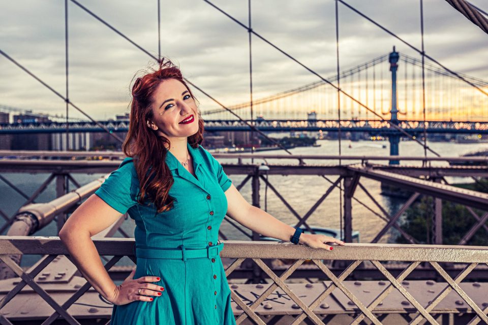 A smiling woman in a turquoise dress leans on a bridge railing.
