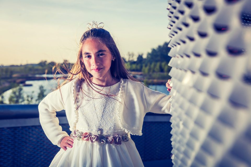 Retrato de una niña con vestido blanco de comunión, cinturón decorado con flores y el cabello recogido.
