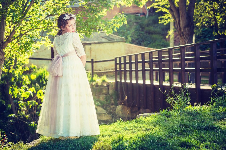 Retrato de una niña con vestido blanco de comunión, fajín rosa y corona floral.