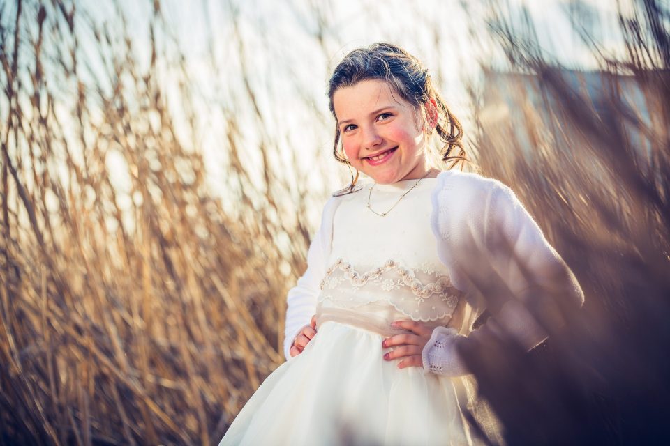 Retrato de una niña con vestido blanco de comunión, sonriente y con las manos en la cintura.