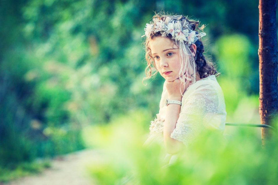Retrato de una niña con vestido blanco de encaje y corona floral, sentada en un parque entre vegetación desenfocada.