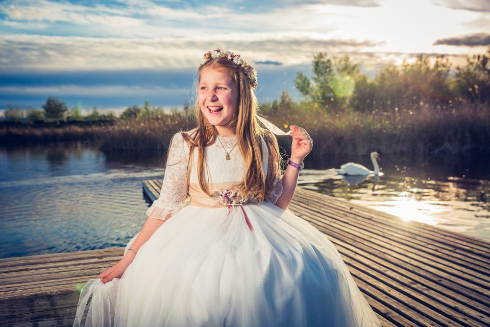 Retrato de una niña con vestido blanco de encaje y una corona floral, sentada en un embarcadero de madera junto al agua.