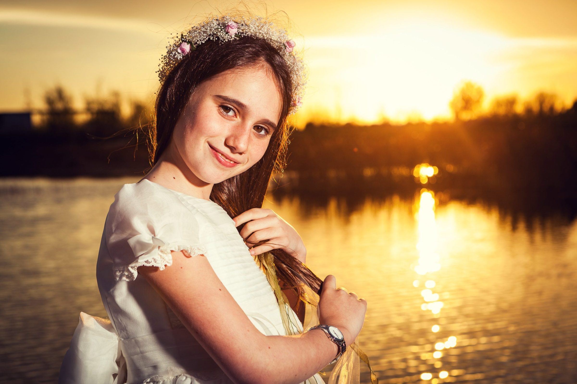 Retrato de una niña con vestido blanco y corona floral, junto a un lago en un parque durante el atardecer.