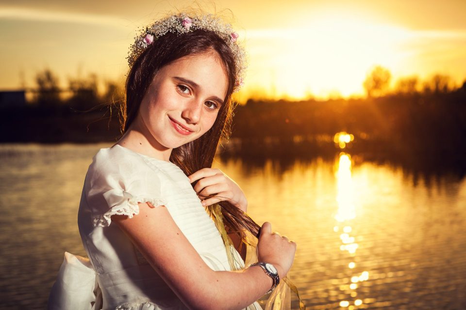 Retrato de una niña con vestido blanco y corona floral, junto a un lago en un parque durante el atardecer.