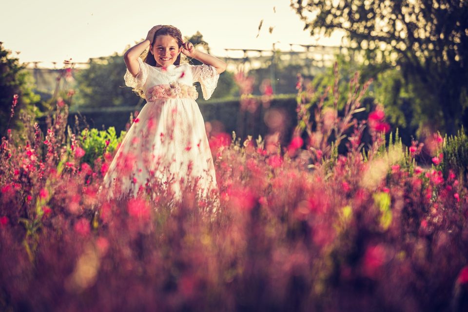Retrato de una niña con vestido blanco y detalles florales, de pie entre flores rosadas en un campo.
