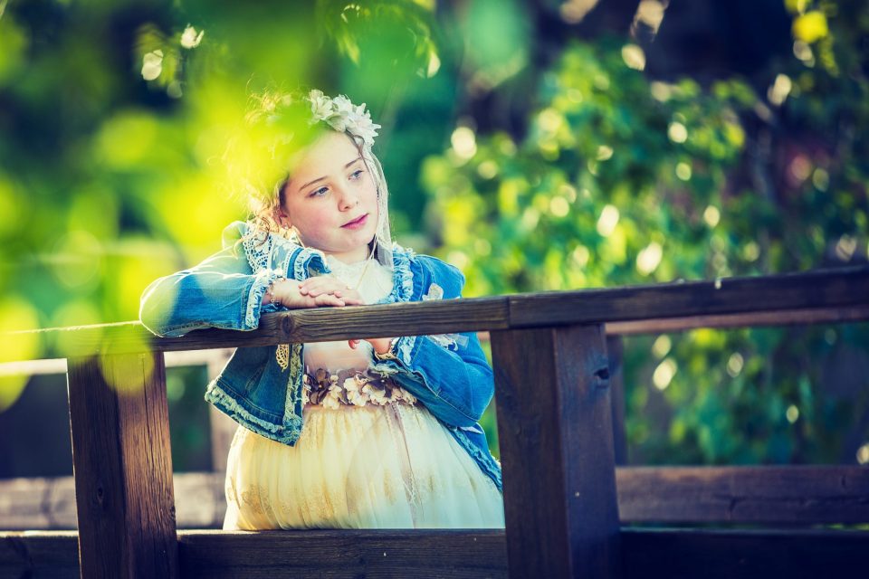Retrato de una niña con vestido claro, chaqueta azul y una corona floral, apoyada en la barandilla de madera de un parque.