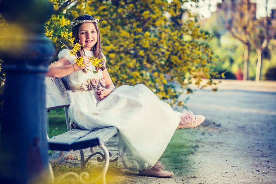 Retrato de una niña sentada en un banco de un parque, con vestido blanco, corona floral y zapatos a juego.
