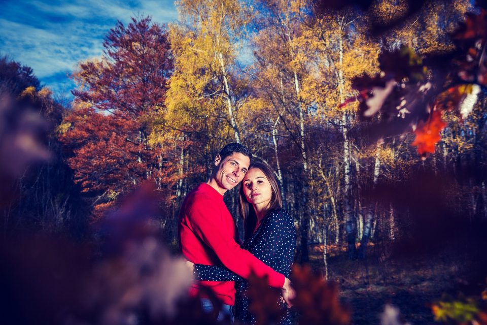 Retrato de una pareja abrazada en un bosque durante el otoño, rodeada de árboles con hojas amarillas, rojizas y anaranjadas.