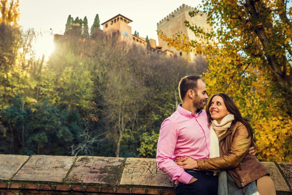 A couple embraces beside a stone wall, surrounded by trees with autumn foliage.