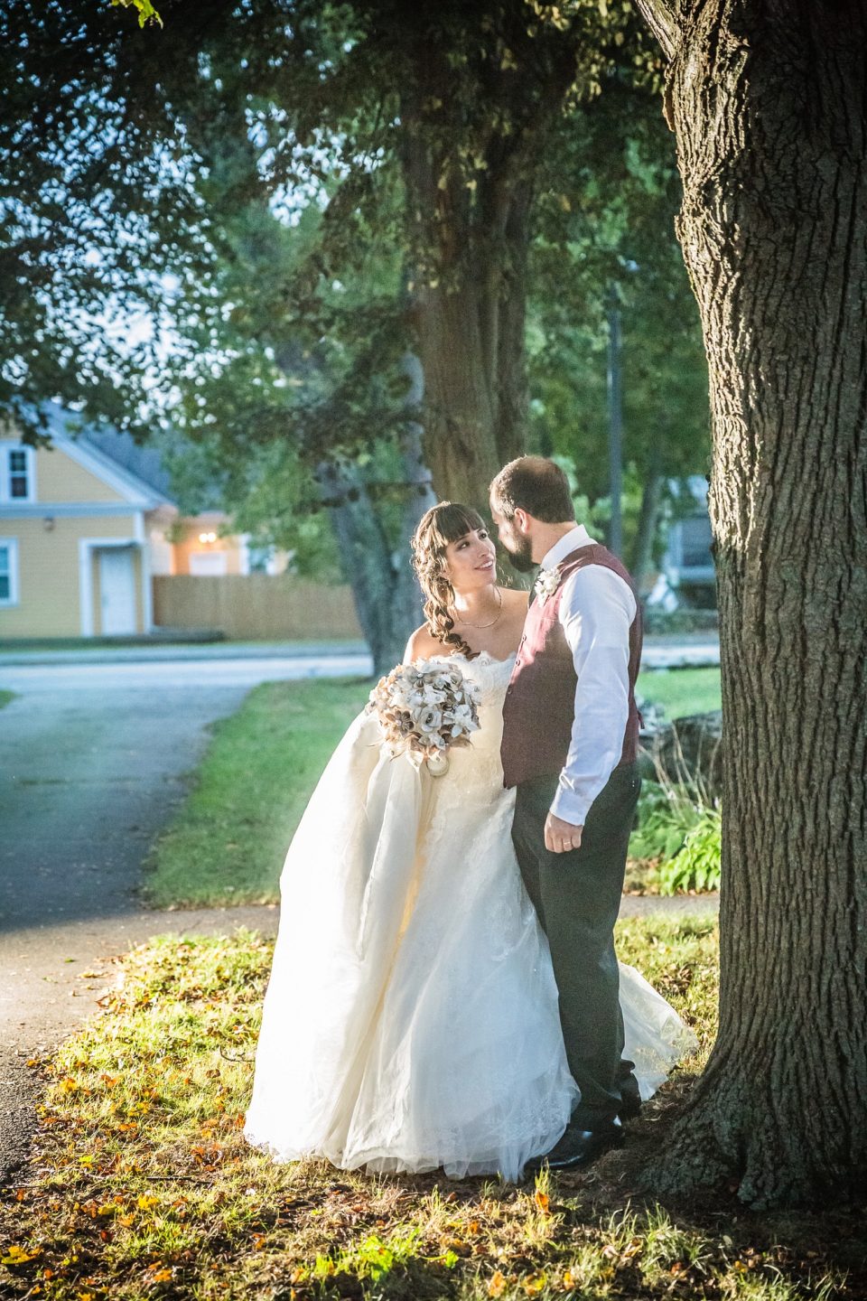A couple in wedding attire pose beside a large tree.