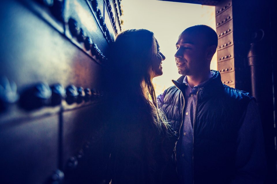 Una pareja se mira de cerca junto a una gran puerta o pared metálica con remaches.