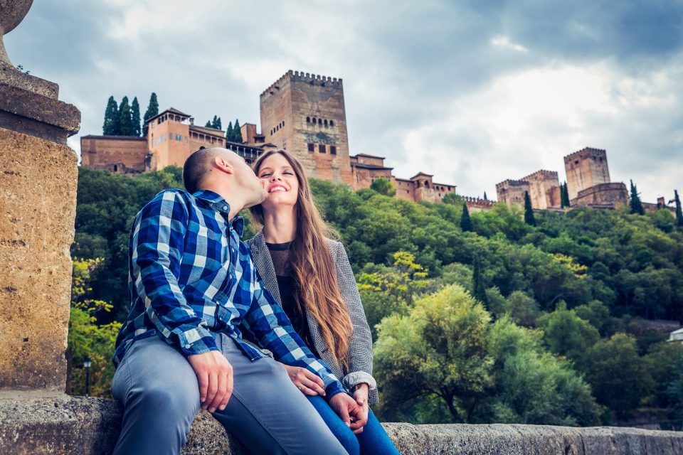 A couple sits in front of a red-hued historic fortress surrounded by lush vegetation.