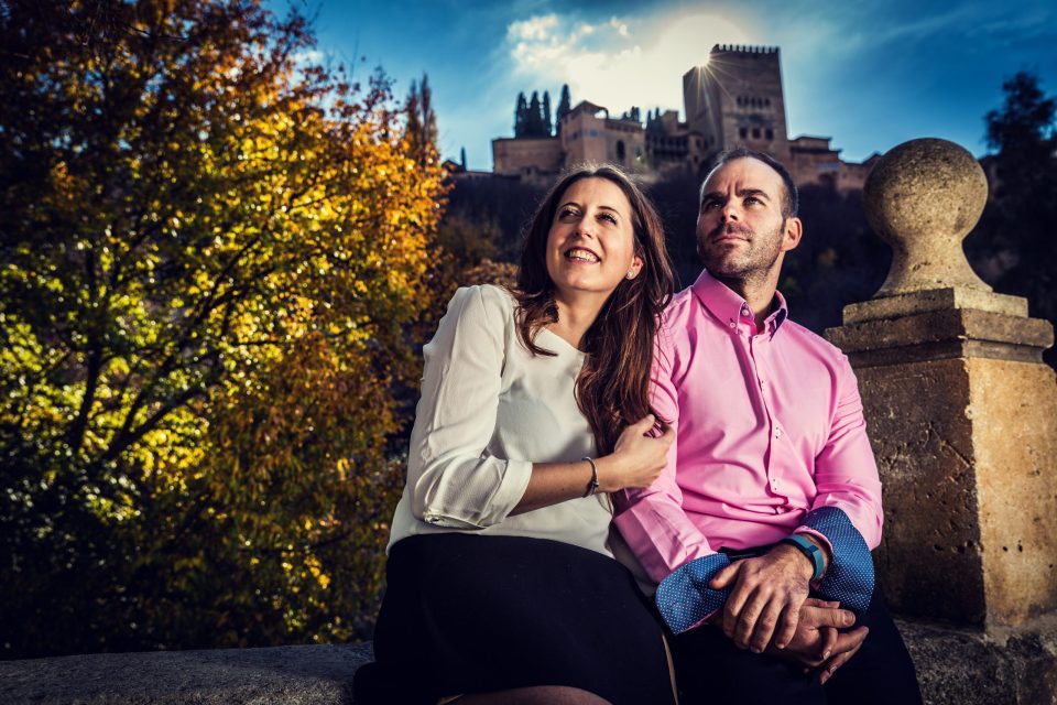 A couple sit beside a stone balustrade, with a historic palace or fortress in the background.