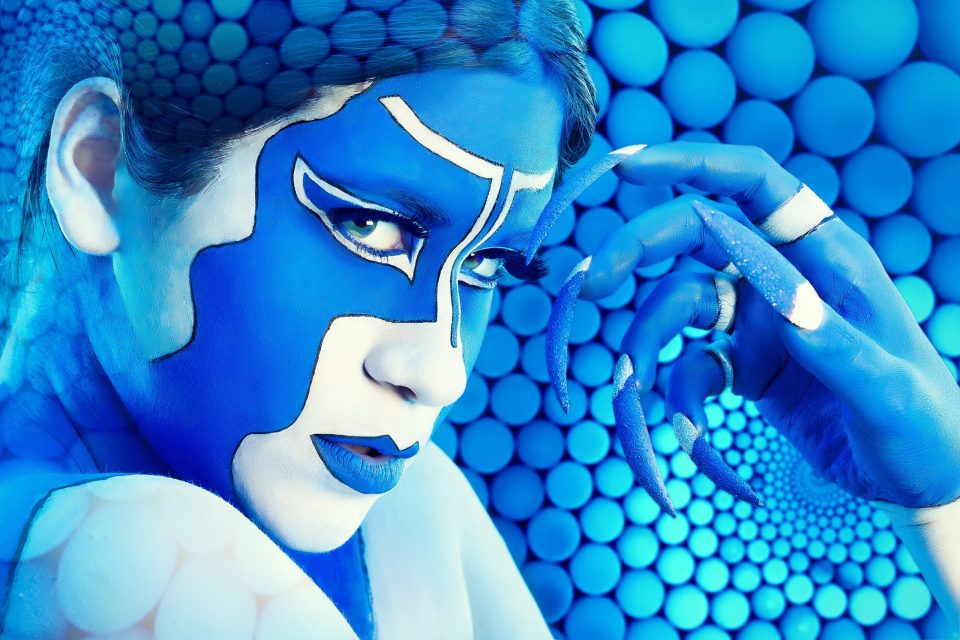 Retrato editorial de fantasía en primer plano de una modelo con el rostro y las manos pintados de azul.