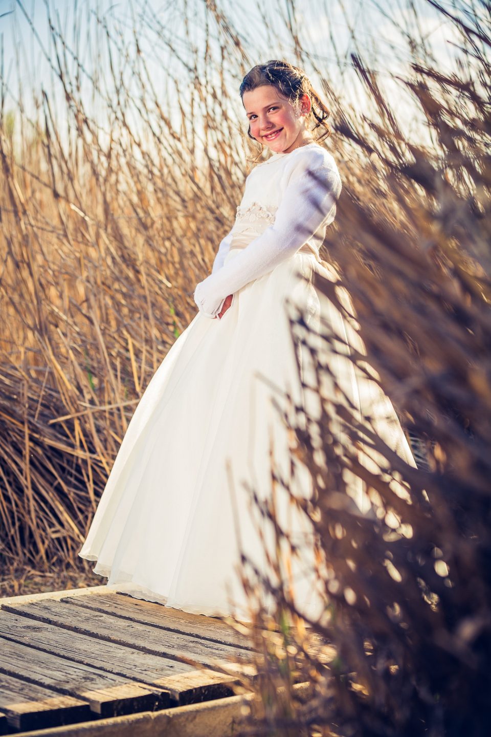 Retrato vertical de una niña con vestido blanco de comunión, sonriente y de pie sobre una pasarela de madera en un parque.