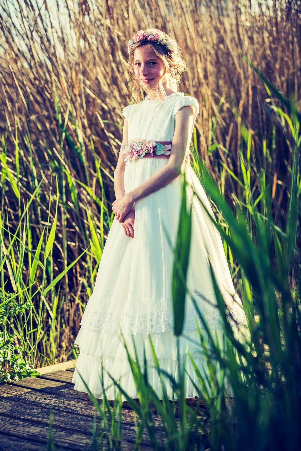 Retrato vertical de una niña con vestido blanco largo, fajín decorado con flores y corona floral.