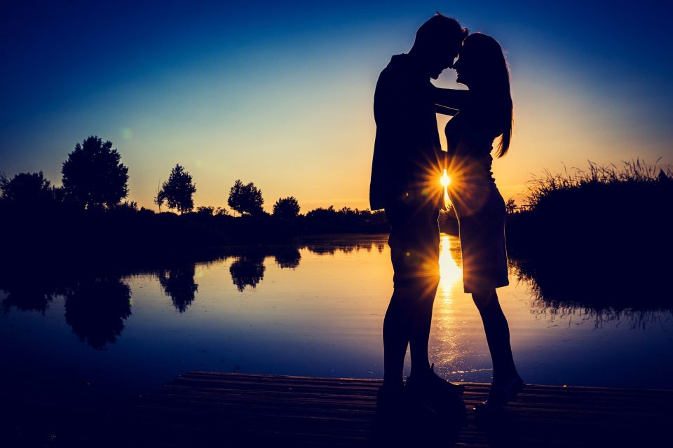 Silueta de una pareja besándose sobre un embarcadero de madera frente a un estanque, en un parque al atardecer.