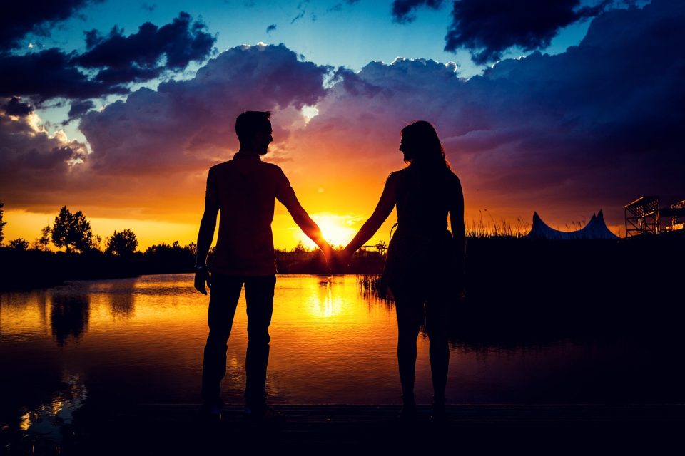 Silueta de una pareja cogida de la mano frente a un lago al atardecer.