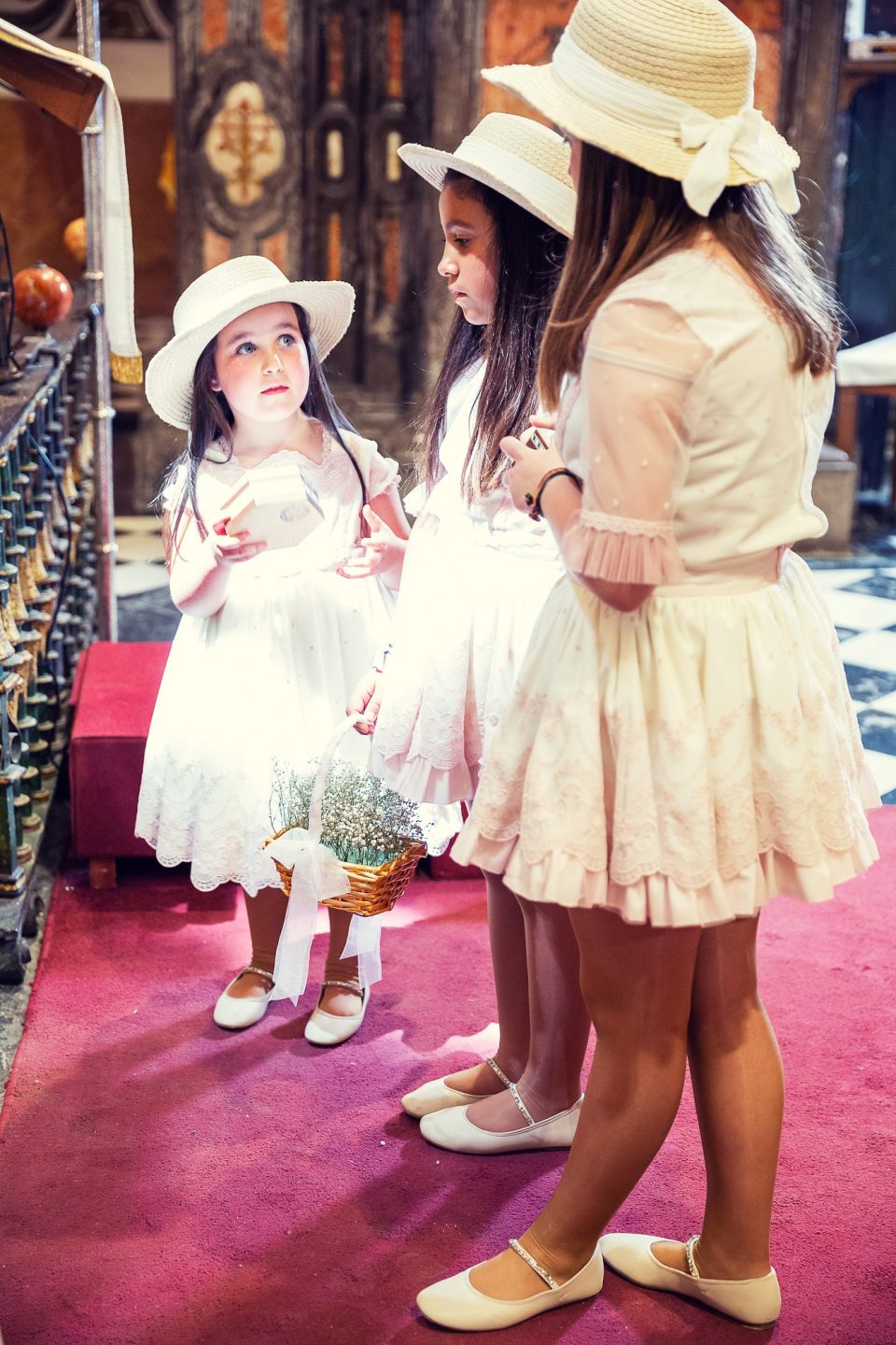 Tres niñas con vestidos claros y sombreros blancos aparecen en el interior de una iglesia, durante una ceremonia de boda.