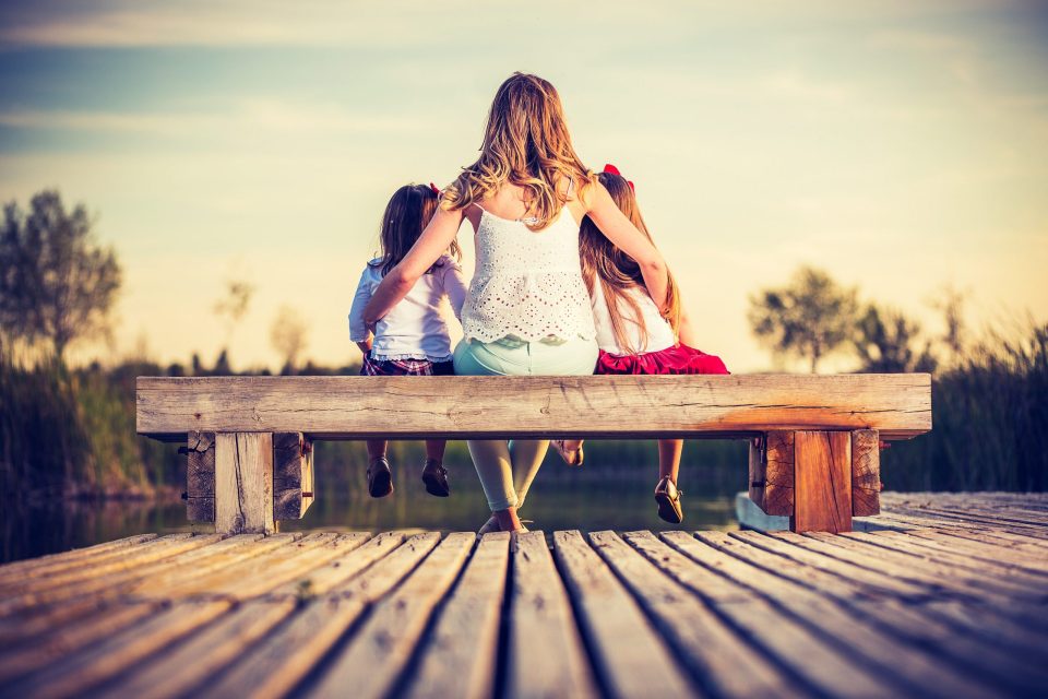 Tres niñas sentadas de espaldas en un banco de madera sobre un muelle, abrazadas entre sí y con las piernas colgando.