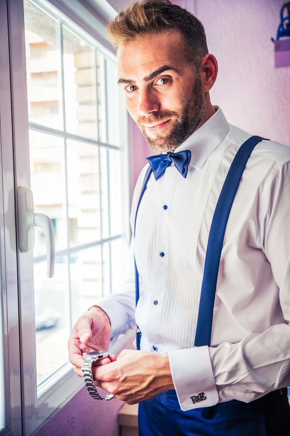 Un hombre con camisa blanca, pajarita azul y tirantes a juego se ajusta el reloj junto a una ventana.