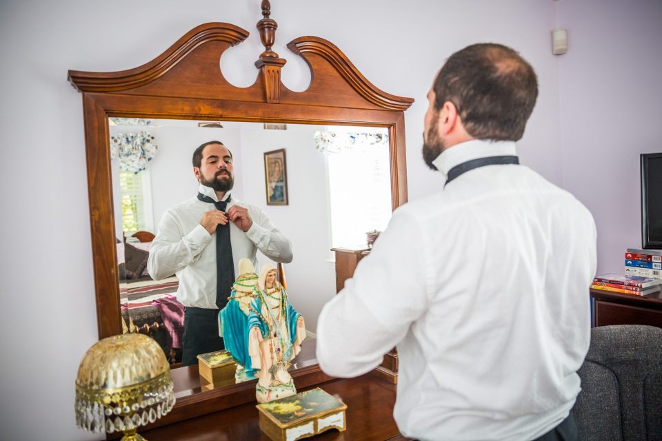 A man in a white shirt and dark tie adjusts his knot in front of a large wooden mirror.