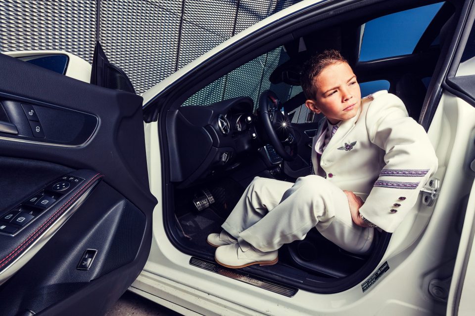 Un niño con traje blanco de ceremonia está sentado en el asiento del conductor de un coche blanco con la puerta abierta.