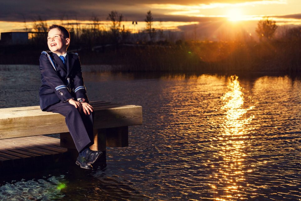Un niño con traje formal oscuro y corbata azul está sentado en un embarcadero de madera sobre el agua.