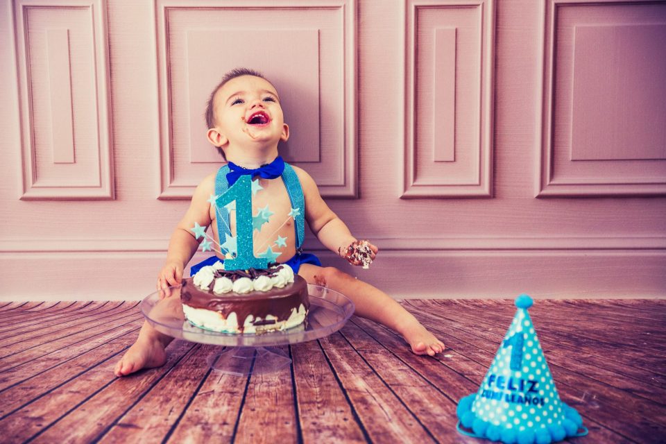 Un niño pequeño sonríe sentado en un decorado de estudio con fondo rosa y suelo de madera.