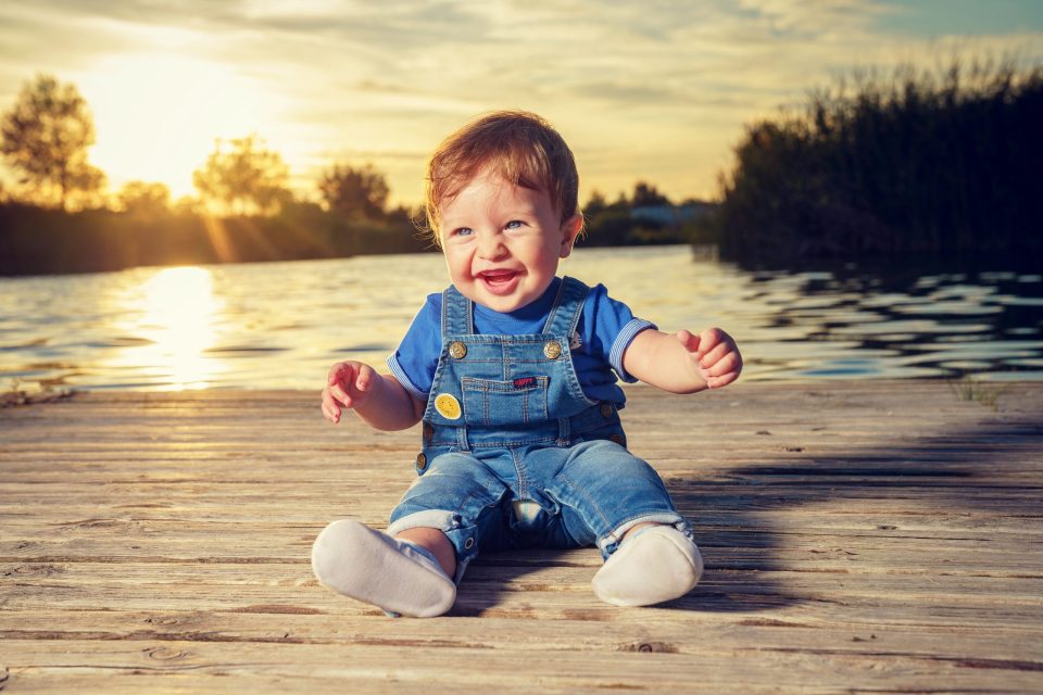 Un niño pequeño sonríe sentado sobre un embarcadero de madera junto al agua.