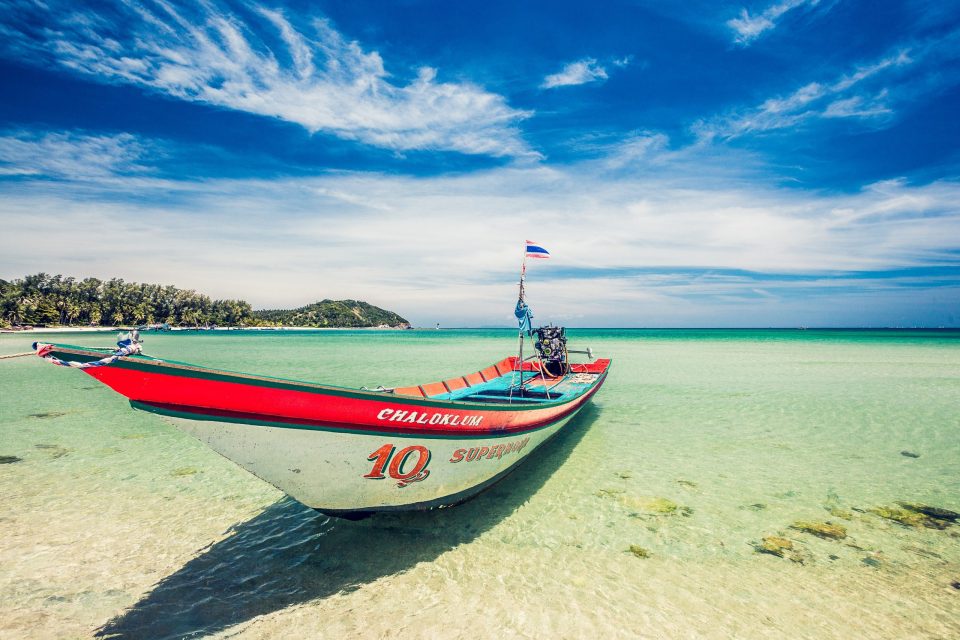 A red and blue longtail boat sits stranded in shallow turquoise waters.