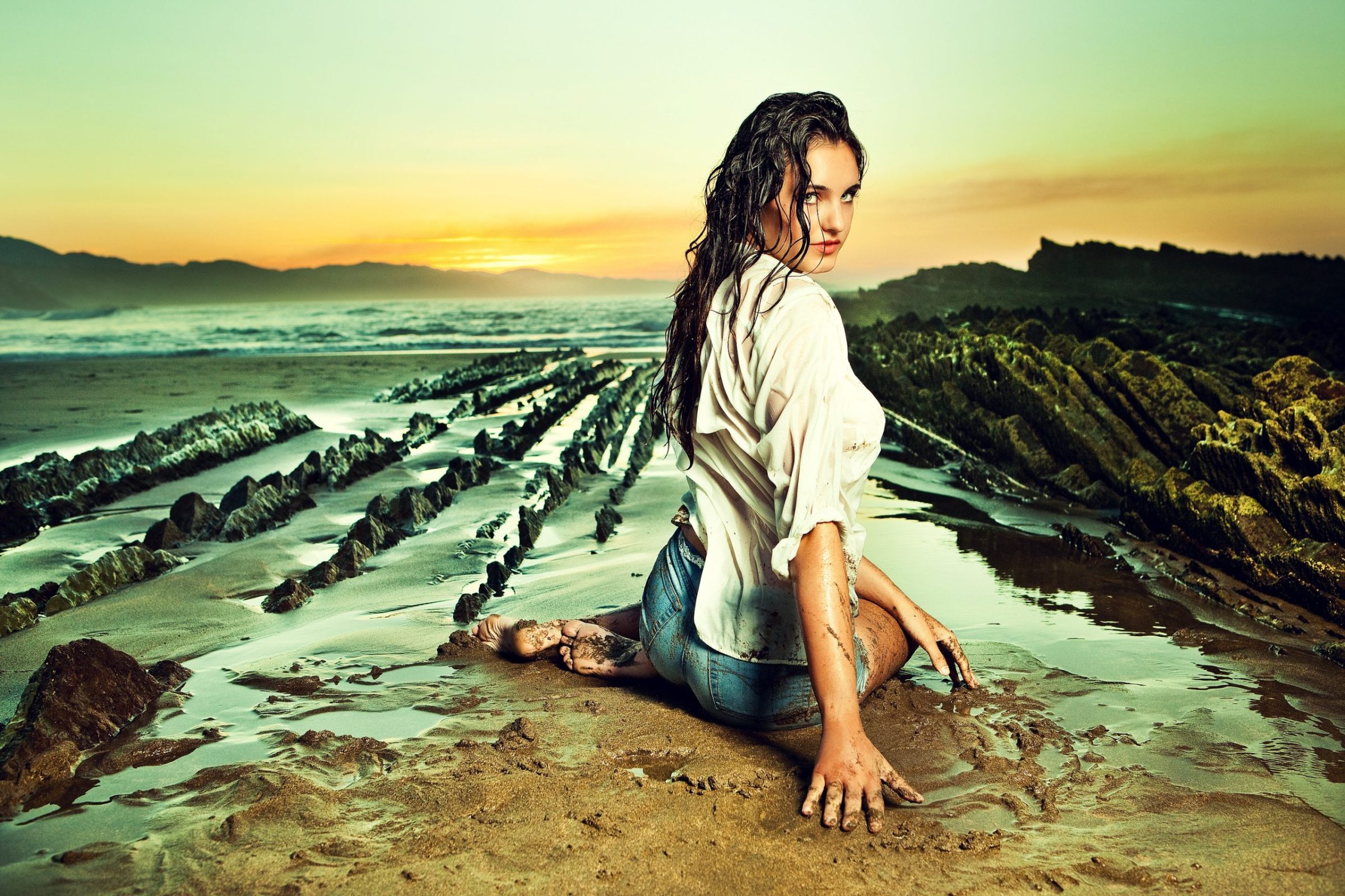 Una modelo con el cabello mojado, camisa blanca y pantalones vaqueros está sentada sobre una zona rocosa de la playa.