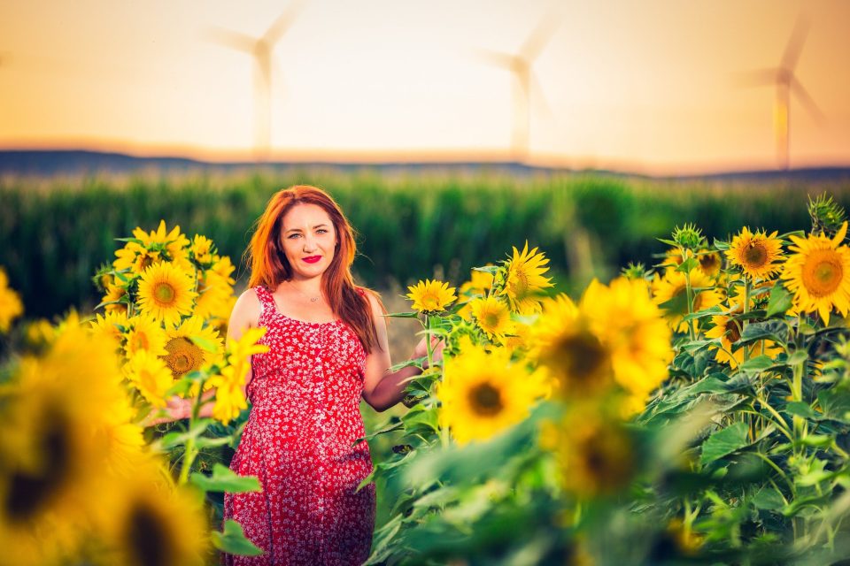 An adult woman in a patterned red dress poses among blooming sunflowers.