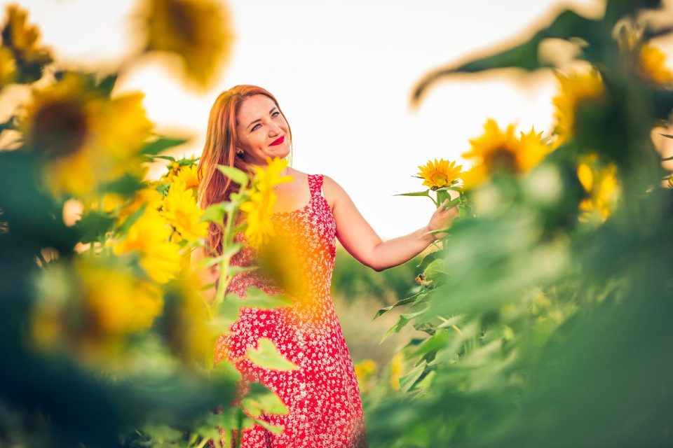 A woman with long reddish hair and a red patterned dress holds a sunflower while posing among several sunflowers.