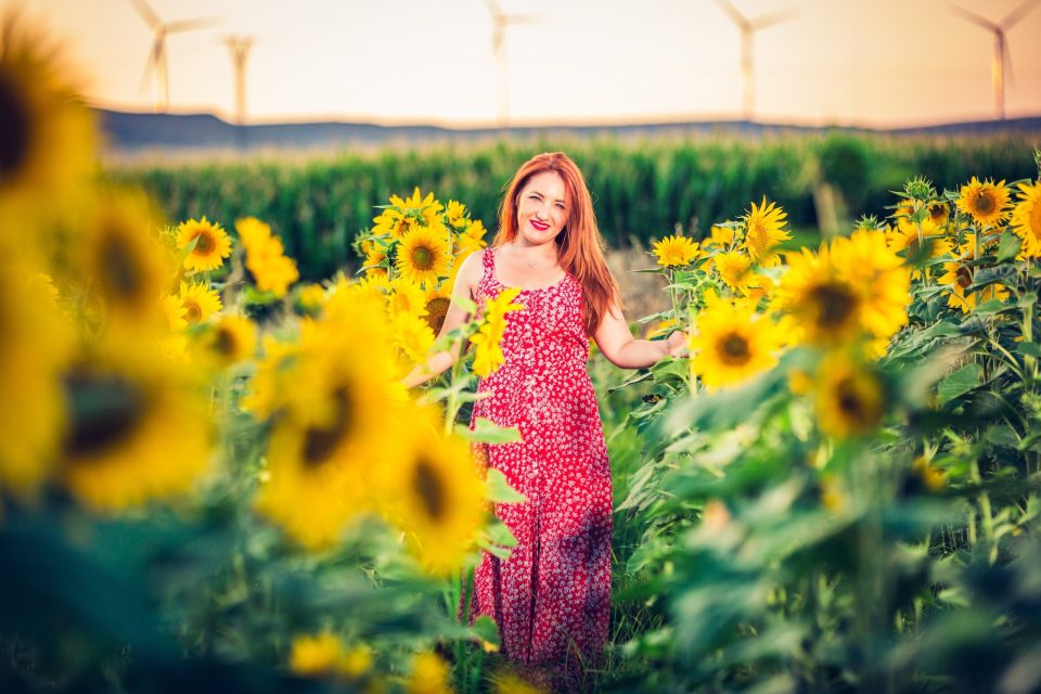 A woman with reddish hair and a red patterned dress poses among tall sunflowers.