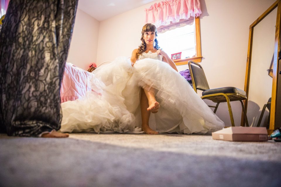 A woman in a voluminous white wedding dress sits posing in a room beside a bed and a chair.