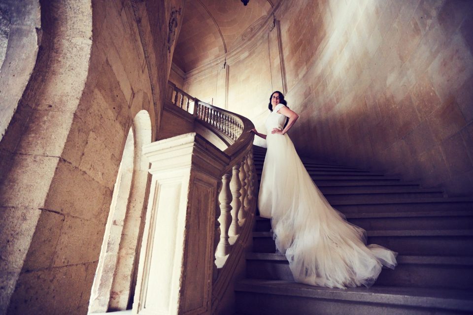 Una mujer con vestido de novia blanco posa en una escalera monumental de piedra.