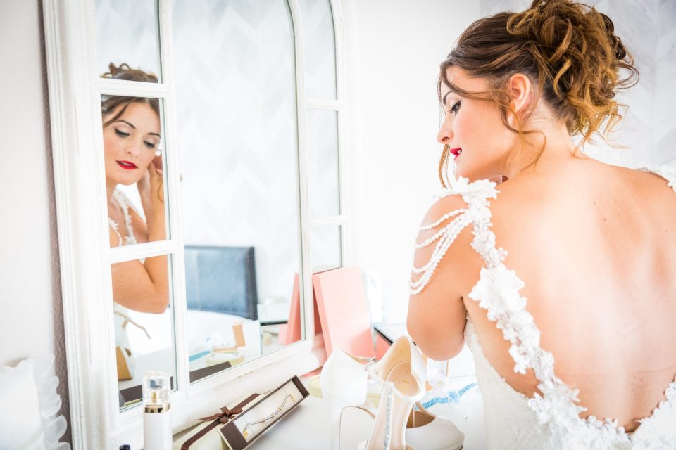 Una mujer con vestido de novia se prepara frente a un espejo en una habitación luminosa.