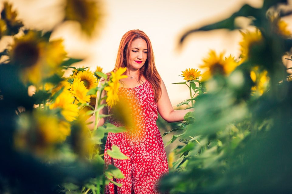 A red-haired woman stands among sunflowers in a red dress patterned with white flowers.