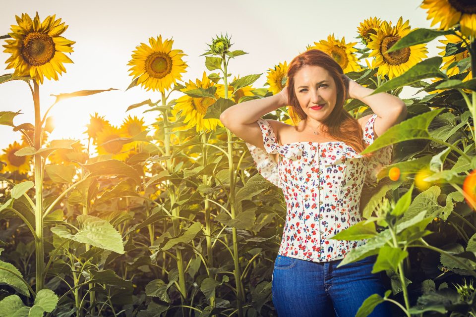 A red-haired woman poses among numerous sunflowers with her arms raised and her hands near her head.