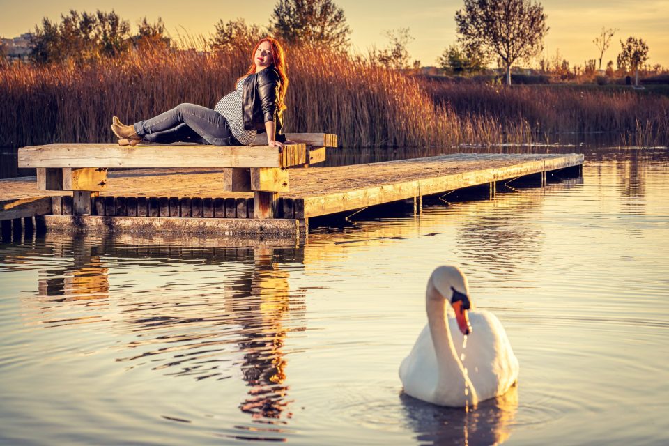 Una mujer embarazada, sentada sobre un muelle de madera, posa junto a un estanque al atardecer.