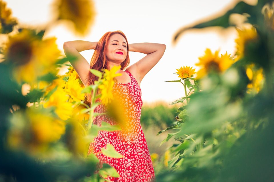 A red-haired woman in a red patterned dress poses among sunflowers.