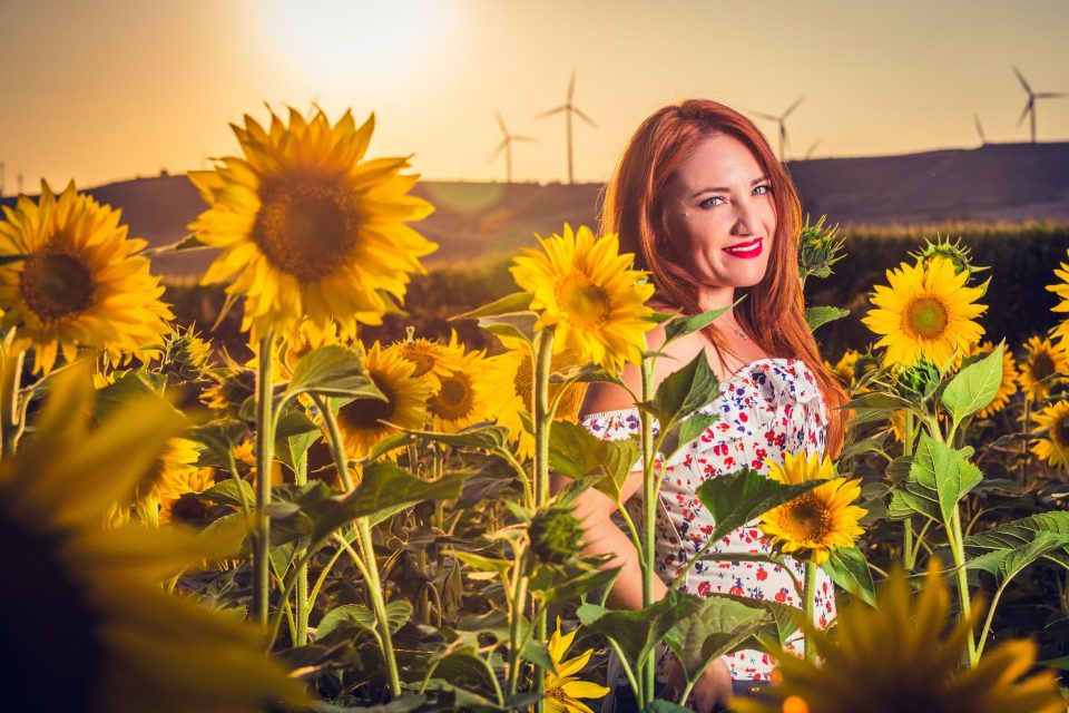 A red-haired woman smiles among tall sunflowers while wearing a white floral-print garment.