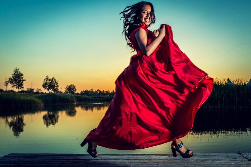 Una mujer sonríe mientras gira y sostiene la amplia falda de un vestido rojo sobre un embarcadero de madera.
