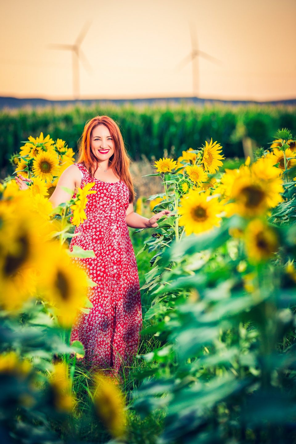 A smiling woman with reddish hair and a patterned red dress poses among blooming sunflowers.