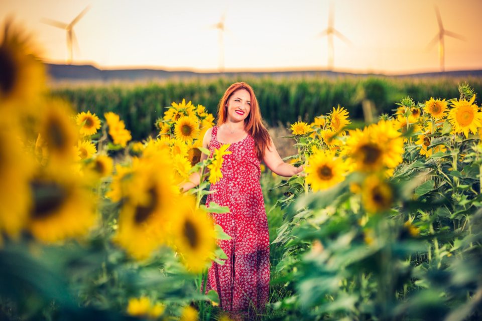 A smiling woman in a red patterned dress poses among blooming sunflowers.