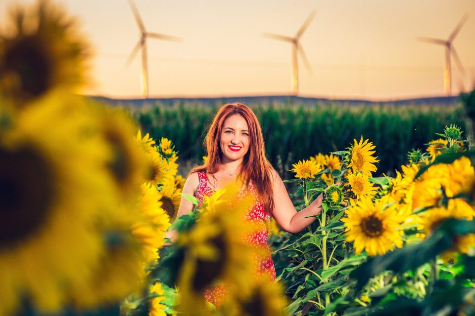 A smiling woman in a red dress stands among flowering sunflowers.